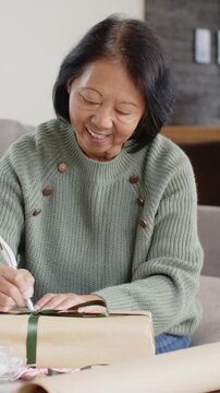 Vertical video: Picking up pen senior Asian woman writing tag, tying ribbon, preparing gift on sofa
