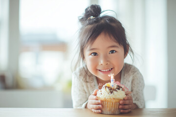 Smiling Asian child holding birthday ice cream with lit candle, sitting at bright kitchen table, joyful moment