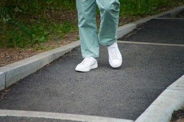 Fototapeta premium Woman’s legs in mint pants walking on asphalt steps in park, wearing white sneakers. Outdoor lifestyle and casual fashion detail. Photo