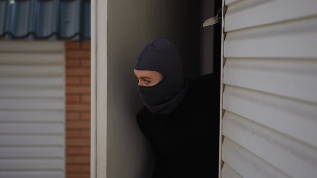 Girl burglar in black clothes and mask carefully moves through garage entry, trying to get inside the house unnoticed.