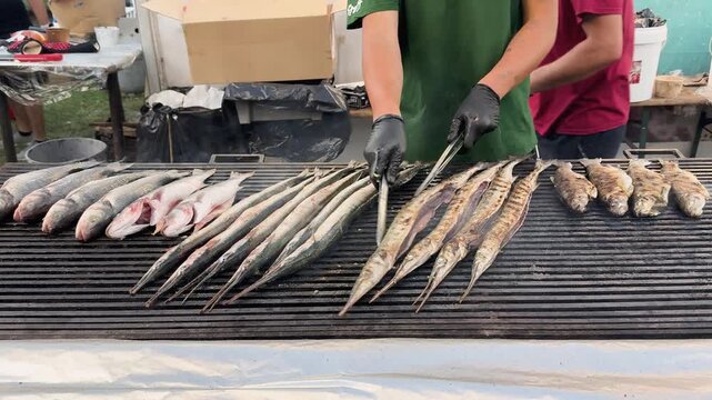 Preparation of Fish on Grill. Hands of chef flipping long whole fish on open grill cooking at outdoor seafood market. Concept of grilling fish, fresh catch, seafood street preparation