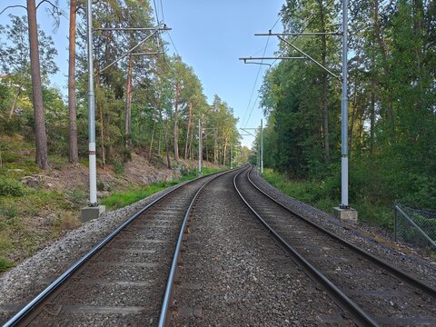 Electrified double railway track curving through green forest in Sweden. Scenic summer view of train and tram infrastructure with catenary masts and gravel ballast.railway in the countryside