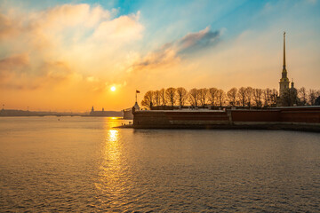 Fototapeta premium Sunset panorama of Peter and Paul Fortress with cathedral spire silhouette and golden sun path on Neva River against warm colored sky. Saint Petersburg, Russia.