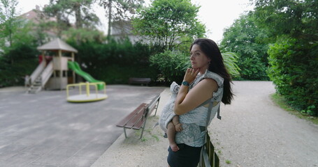 Mother with baby in a carrier standing in a park, gazing into the distance with a thoughtful expression, enjoying a calm moment of reflection amidst nature