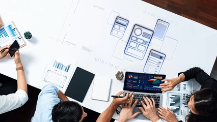 A diverse team engages in collaboration around a modern workspace, focusing on digital product design and prototyping with laptops and mockups visible. Raster