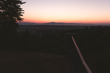 Beautiful sunrise over countryside with soft pastel sky, peaceful landscape viewed from balcony railing.