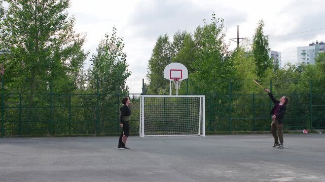 Two People playing shuttlecock kicking game on a grey concrete court in an outdoor park area during daylight