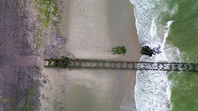 Aerial view of ocean waves crashing against rows of stone breakwaters. The geometric pattern of the structures contrasts with the dynamic movement of the sea, creating a striking coastal scene.