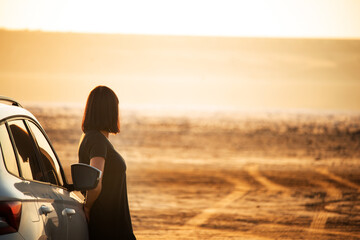 Woman in black dress leaning on car and looking at the horizon on a sandy road. Concept of travel,...