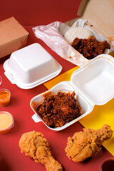 Foam clamshell filled with hot sauced fried chicken, plus drumsticks and dips on a bold red–yellow background; spicy fast-food takeaway/delivery menu shot.