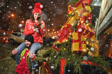 woman sits on the shoulders of a bearded man and smiles against the backdrop of a red retro car with Christmas gifts on the roof and against the backdrop of snow