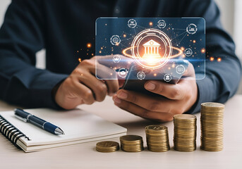 Person using smartphone with glowing bank icon and financial icons, stacks of coins, and notebook on table