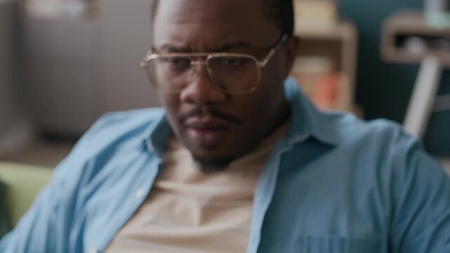 Tilt up of tense African American man wearing eyeglasses and casual attire switching TV set while pressing power button on remote control, watching broadcast and expressing dissatisfaction