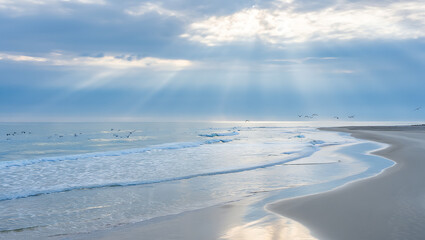 Sunbeams pierce through dramatic clouds over a serene ocean, illuminating a tranquil beach with gentle waves and distant seabirds in flight