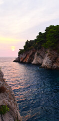 Dramatic rocky coastline with layered cliffs and lush green pines at sunset, overlooking calm blue sea waters reflecting soft pink and orange hues.