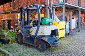 Old forklift with gas cylinder parked in an industrial yard near a brick building. Industrial equipment and machinery used for cargo handling and transportation.