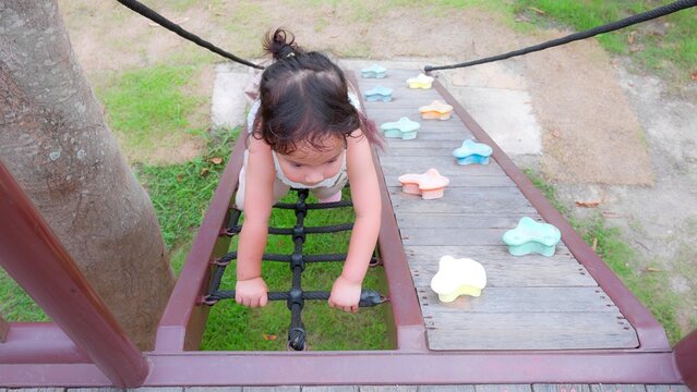Happy Young Asian child girl climbing a rope bridge at a playground, showcasing outdoor play and adventure.