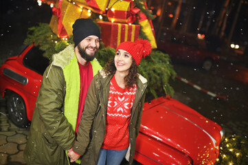 couple of lovers - a man and a woman hold hands and smile against the background of a red retro car with Christmas gifts on the roof