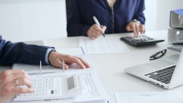 Two professional accountants collaborating in a modern office, calculating taxes and analyzing financial data with calculators, documents, and a laptop. Audit and taxes in business
