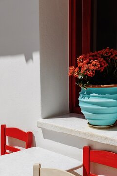 Charming detail of a traditional Cycladic house in Apiranthos village on Naxos island, Greece, featuring bright red chairs, a turquoise flower pot with blooming flowers on a whitewashed windowsill.