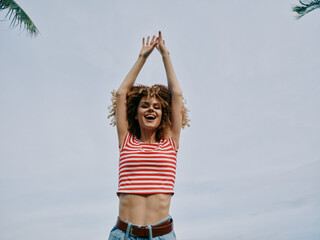 Vibrant portrait of a joyful woman raising arms beneath a clear sky, wearing a red and white...