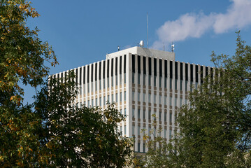 Upward view of a modern office tower with vertical black stripes on the roofline, framed by green...
