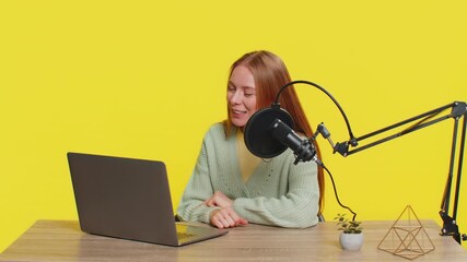 Happy excited woman blogger at table recording podcast, giving workshop, webinar. Adult girl influencer speaking into microphone and laptop webcam, making video call, discussing on yellow background.