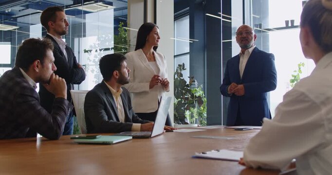 Business meeting with corporate strategy presentation. Leader explaining financial plan to manager team sitting and standing at table in glass office, discussion of goals and next steps by colleagues