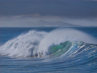 Mighty ocean wave peels with a thick crest of white foam and offshore spray. Sunlight illuminates the water, revealing a vibrant emerald green core that stands out against the dark blue curling wave.