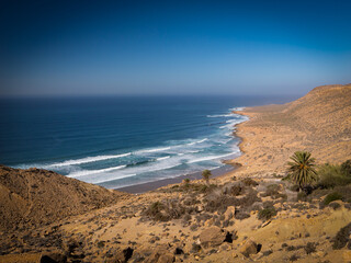 Secluded sandy beach with rolling waves breaking along the rugged coastline. Peaceful coastal landscape of Morocco with turquoise water and the surrounding arid hills create a stunning color contrast.