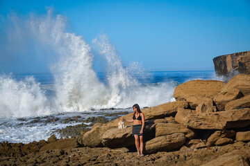 PORTRAIT: Beautiful woman in dark skirt and bikini top cuddles a white cat on a rocky beach as...
