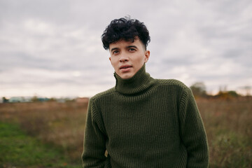 A candid portrait of a young man with curly hair wearing a thick olive sweater, conveying...