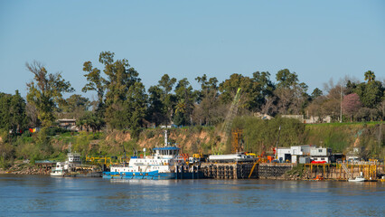 Fototapeta premium River scene featuring tugboats and industrial activity along a waterfront. Represents shipping, transport, construction and port operations in a natural setting