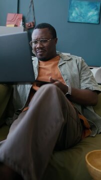 Vertical shot of young anxious African American man in casual attire sitting on couch in living room and looking at laptop screen while watching match broadcast
