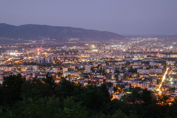 long-exposure shot of the Skopje cityscape at twilight, with the city lights sparkling against the backdrop of the surrounding mountains and a hazy sky