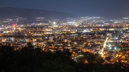 panoramic view of the Skopje cityscape at night, showcasing the illuminated urban landscape with a mountain range in the background. The glowing city lights