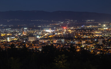 panoramic night view of a sprawling cityscape, with the illuminated buildings and streets of Skopje nestled against the backdrop of mountains