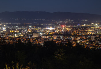A panoramic, nighttime view of a vibrant cityscape. The city lights illuminate the urban landscape against the dark sky and mountains