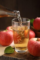 Pouring delicious apple cider from bottle into glass at wooden table against dark background, closeup