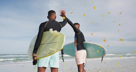 High-fiving surfers holding surfboards with leashes on sandy beach facing ocean under cloudy sky