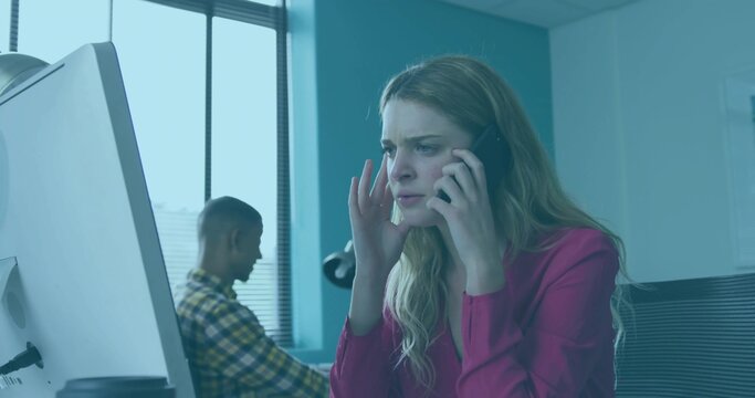 Talking woman in magenta blouse touching temple, speaking on smartphone in office with monitor