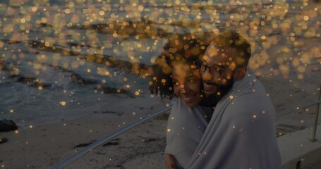 Leaning against metal railing at seaside promenade, woman and man wrapping in light-colored blanket