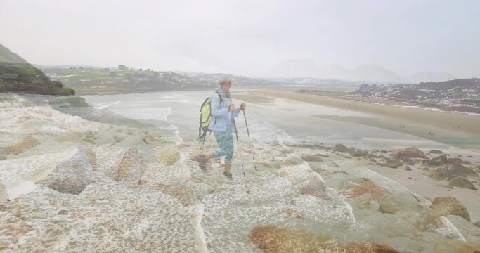 Balancing senior hiker on wet coastal rocks, with backpack, trekking poles and boots