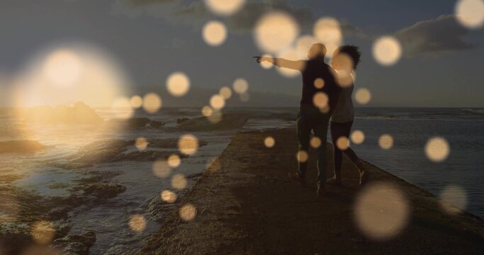 Walking couple pointing toward ocean rocks at sunset with bokeh orbs on stone pier, copy space
