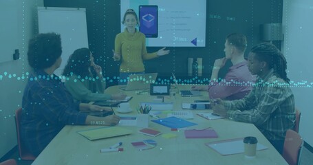 Presenting woman in mustard top gesturing toward screen in meeting room with laptops and charts