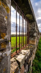 A weathered stone wall with a rusty metal fence, showcasing a view of a grassy field and a cloudy sky.