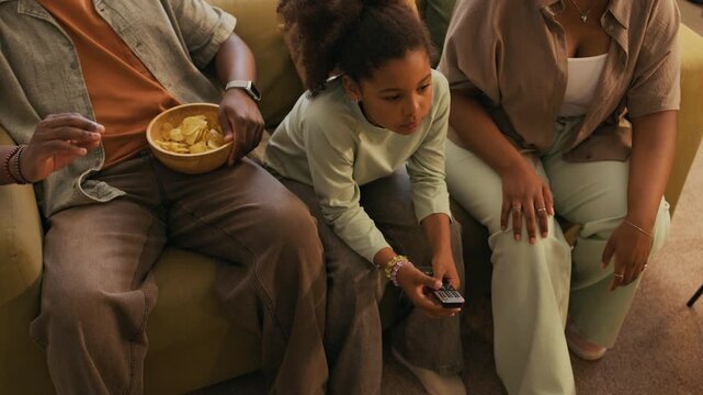 High angle of cute youthful girl with remote control switching channels while sitting on couch between her parents and spending pastime together watching and discussing stuff on TV