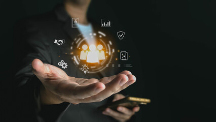 Businessman holding a smartphone and projecting a glowing hologram of business management icons. Concepts like teamwork, strategy, and security are shown over his open palm.