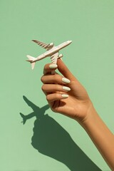 A hand holds a small white airplane against a mint green background.  The shadow of the plane is cast on the background.  Manicured nails are a light mint green color