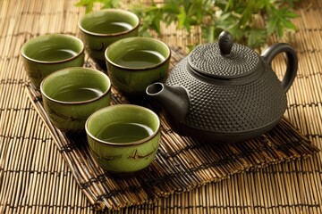 A traditional tea set with four small green teacups and a dark, textured teapot on a woven bamboo mat.  Green tea is poured into each cup